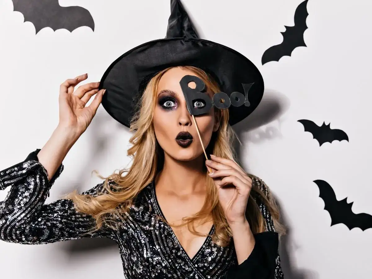 Kobieta w kapeluszu czarownicy z literami "Boo!" przed twarzą. Jej prosty makijaż czarownicy podkreśla oczy i usta.