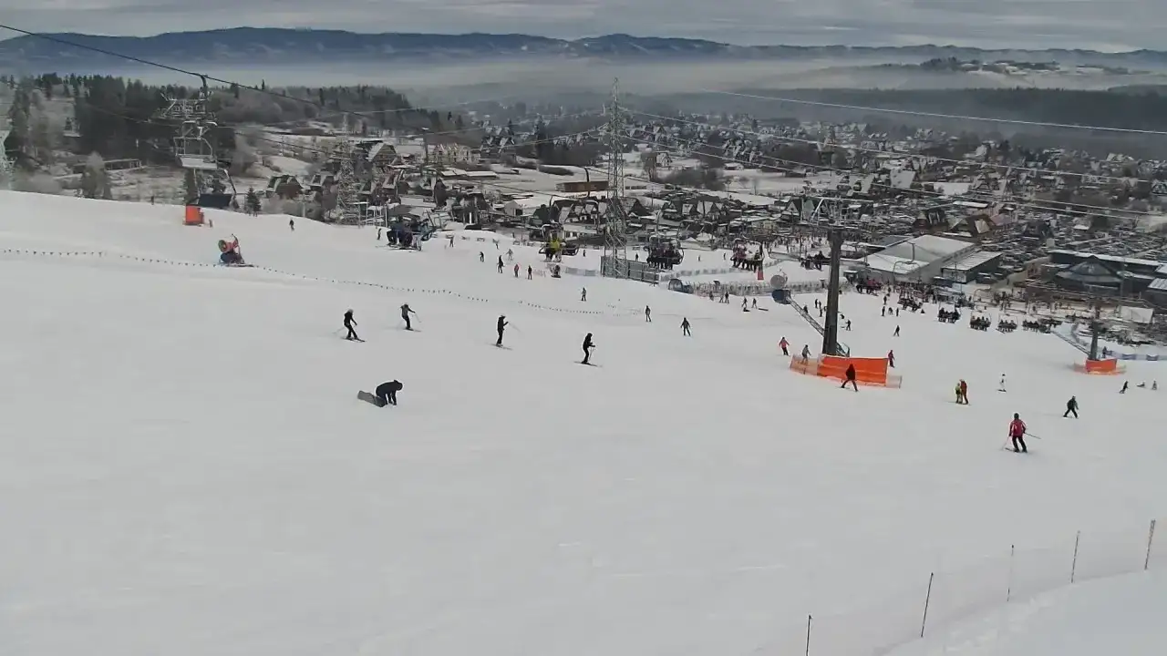 Widok z kamery na stok narciarski w Bodzentynie. Narciarze i snowboardziści szusują po ośnieżonych zboczach, w tle widoczne zabudowania i góry.