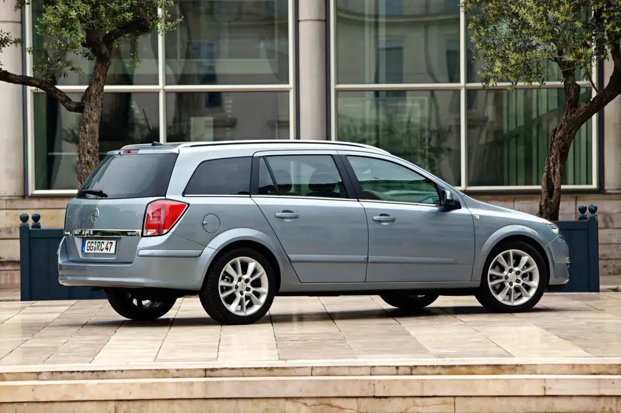 Srebrny Opel Astra 2005 kombi zaparkowany przed nowoczesnym budynkiem z drzewami.