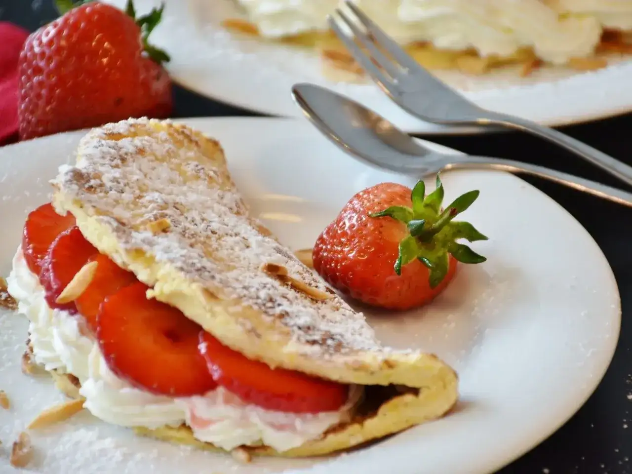 Puszysty omlet z bitą śmietaną i truskawkami, posypany cukrem pudrem i płatkami migdałów. Obok leży świeża truskawka.