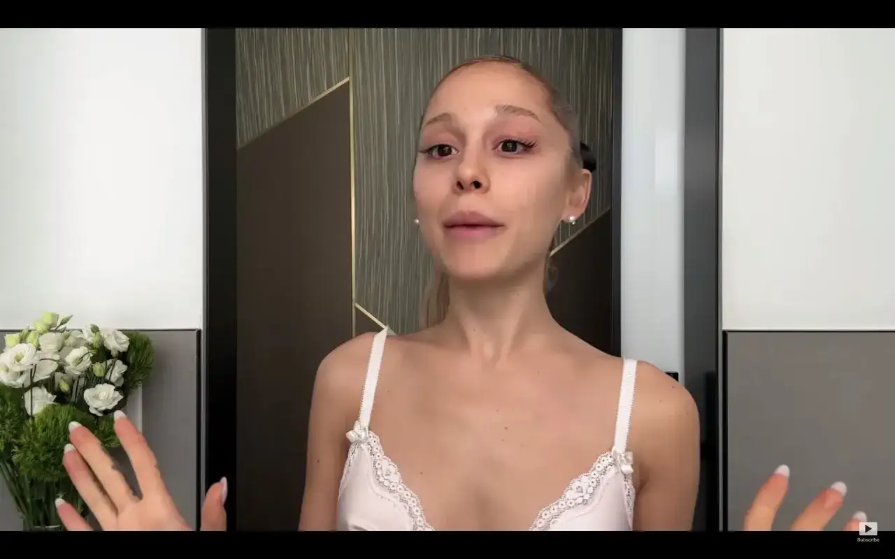 Ariana Grande bez makijażu, w białej koronkowej bieliźnie, z bukietem białych kwiatów po lewej stronie.