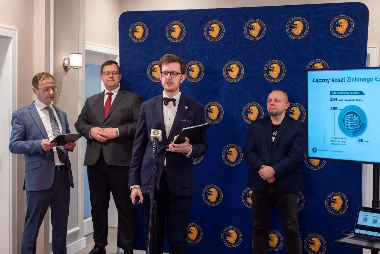 Czterech mężczyzn w garniturach stoi przed niebieskim tłem z logo. Jeden z nich mówi do mikrofonu.