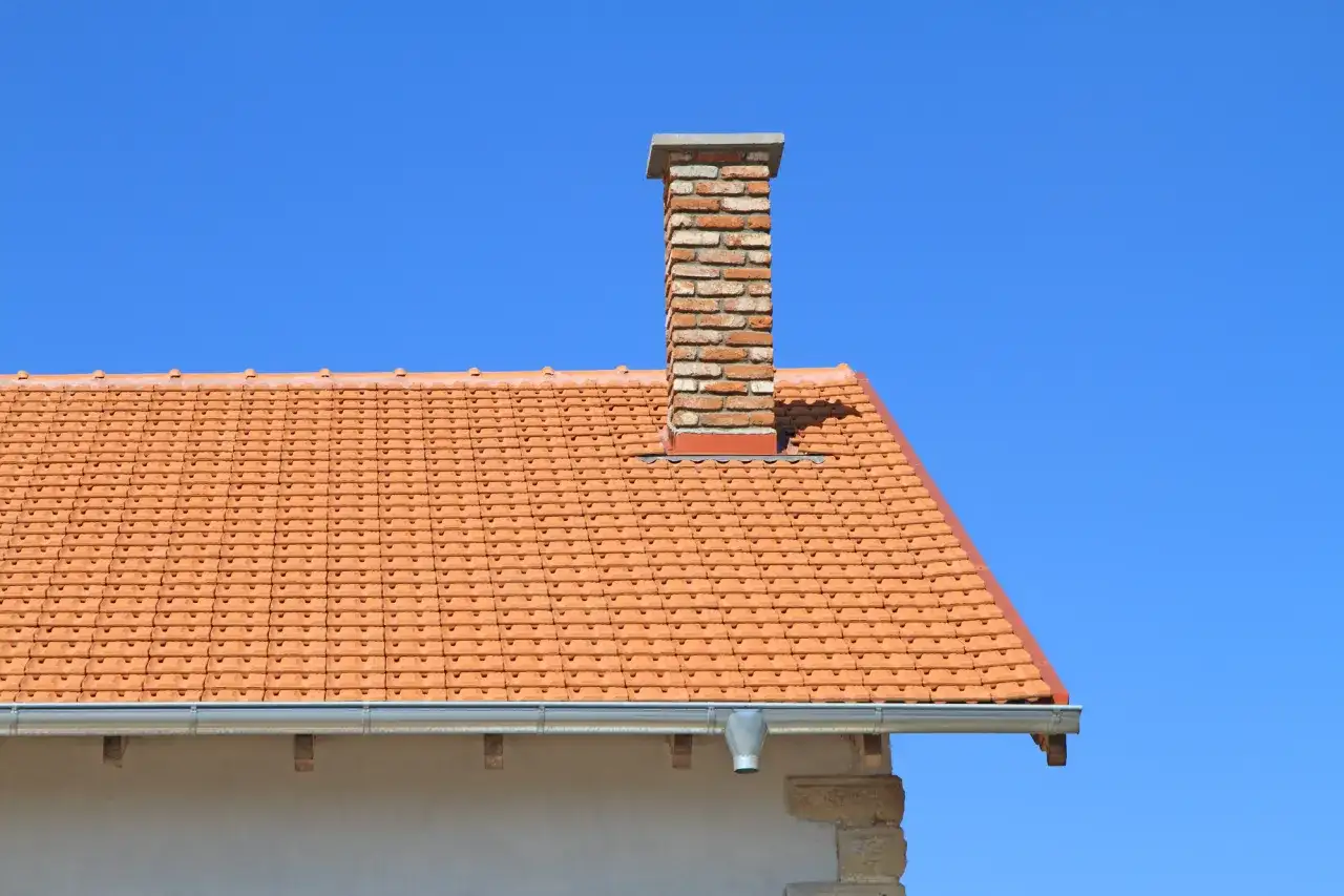 Ceglany komin na dachu z pomarańczowej dachówki. Zastanawiasz się, ile kosztuje budowa komina?
