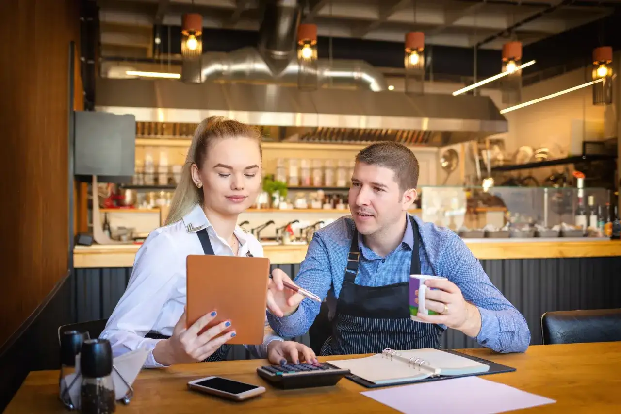 Para omawia koszty uzyskania przychodu w restauracji, analizując dane na tablecie i kalkulatorze.