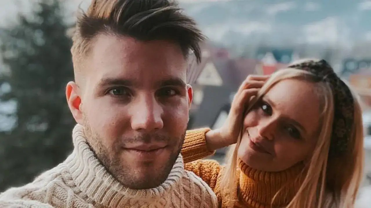 Olga Kalicka i jej partner w ciepłych swetrach, uśmiechnięci na tle górskiego krajobrazu.