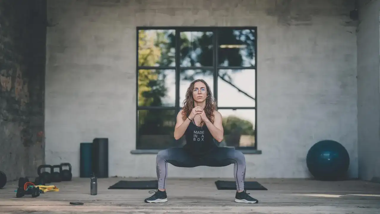 Kobieta wykonuje przysiad sumo. Widoczne hantle, piłka gimnastyczna i maty. Trening HIIT efekty widać po jej sylwetce.