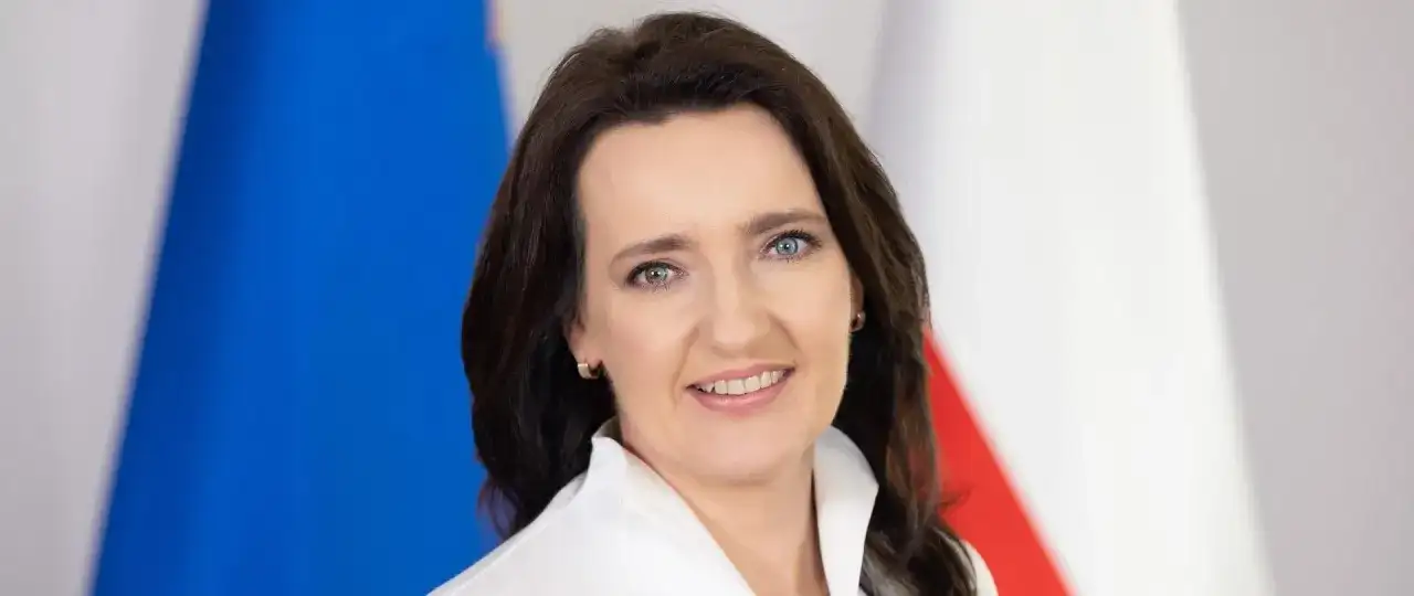 Marzena Okła-Drałowicz, uśmiechnięta kobieta z ciemnymi włosami, na tle biało-czerwonej flagi.