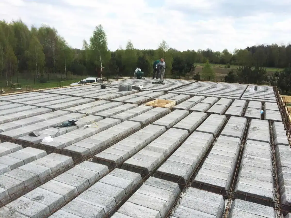 Budowa stropu teriva w toku. Betonowe pustaki ułożone w rzędach, zbrojenie gotowe do zalania. Pracownicy doglądają postępu prac.