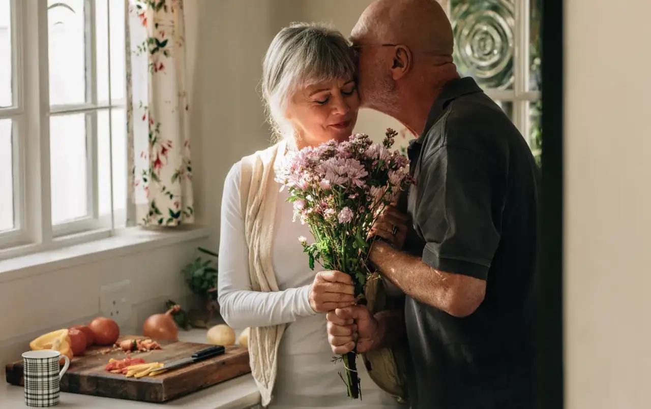Starszy mężczyzna całuje w czoło kobietę trzymającą bukiet kwiatów.