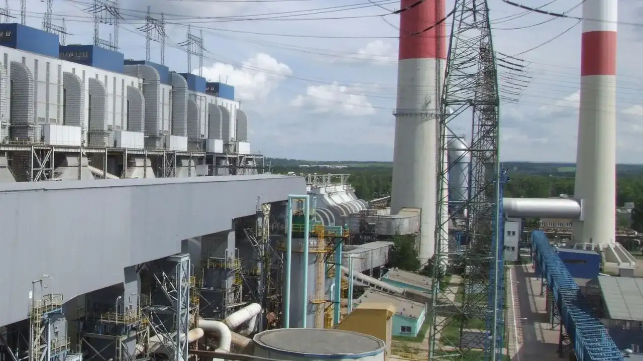 Duże budynki elektrowni z niebieskimi elementami, wysokie kominy z czerwonymi pasami i linie energetyczne.