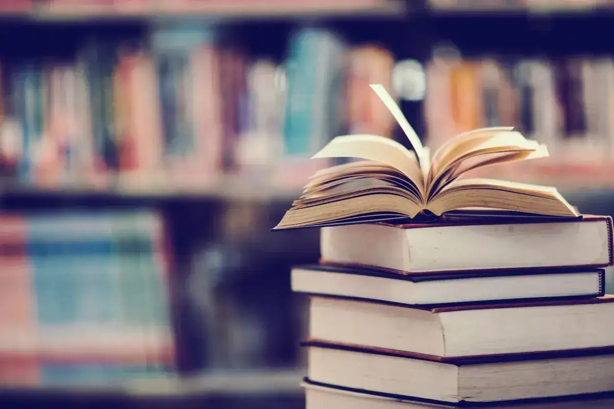 Stos książek, na wierzchu otwarta księga. W tle regały pełne literatury, w tym podręczniki do prawa pracy.