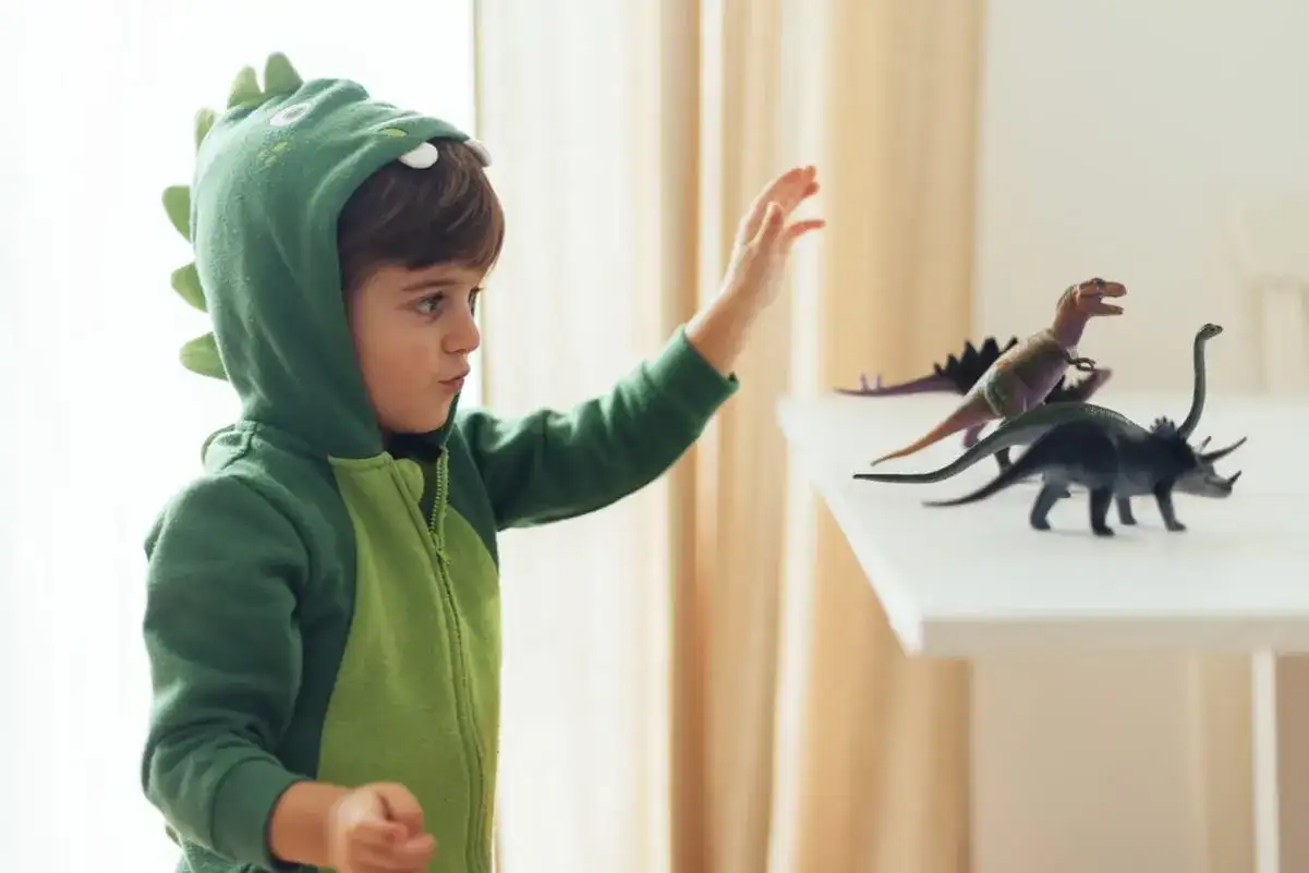 Chłopiec w zielonym stroju dinozaura DIY sięga po figurki prehistorycznych gadów.