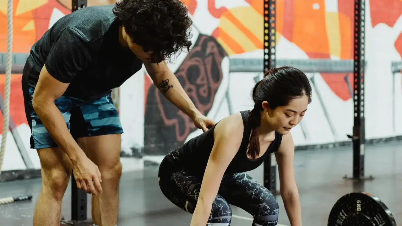 Entrenador ayuda a mujer a levantar pesas en gimnasio con mural colorido de fondo.
