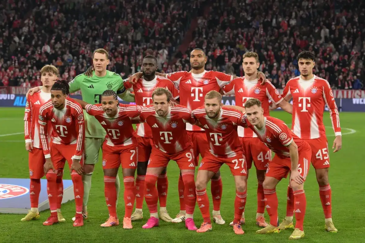 Drużyna Bayern Monachium – zawodnicy w czerwono-białych strojach pozują do zdjęcia na murawie stadionu, z trybunami pełnymi kibiców w tle.