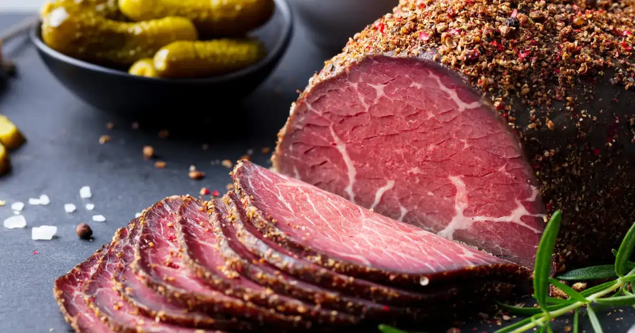 Pokrojone plastry pastrami, przyprawione pieprzem, obok ogórków konserwowych. Zastanawiasz się pastrami co to? To aromatyczne, peklowane i wędzone mięso wołowe.