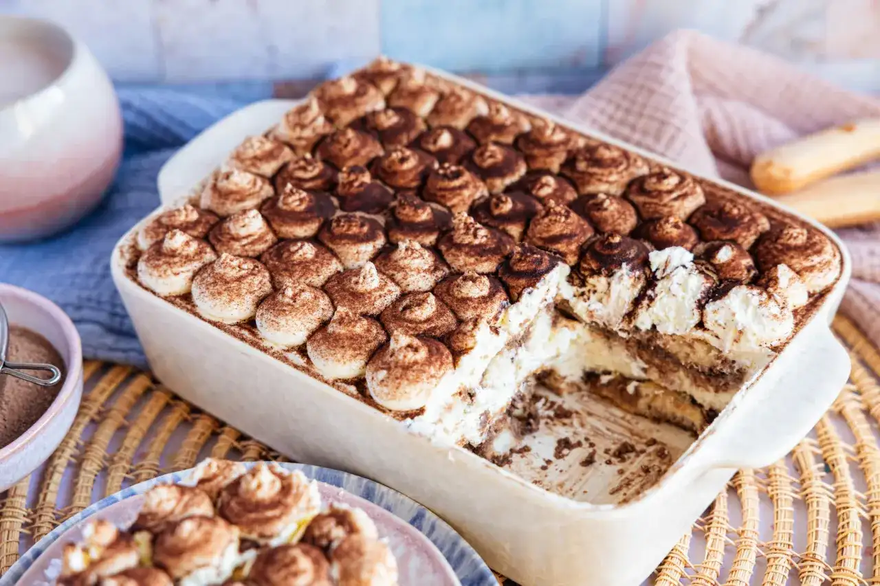 Cremiges, veganes Tiramisu ohne Alkohol, bestäubt mit Kakao. Ein Stück wurde bereits entnommen, was die Schichten aus Creme und Löffelbiskuits zeigt.