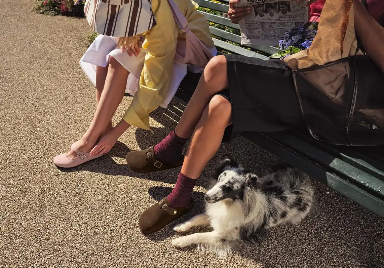 Dwie kobiety siedzą na ławce, prezentując modne buty na lato: różowe baleriny i brązowe klapki. Obok leży pies.