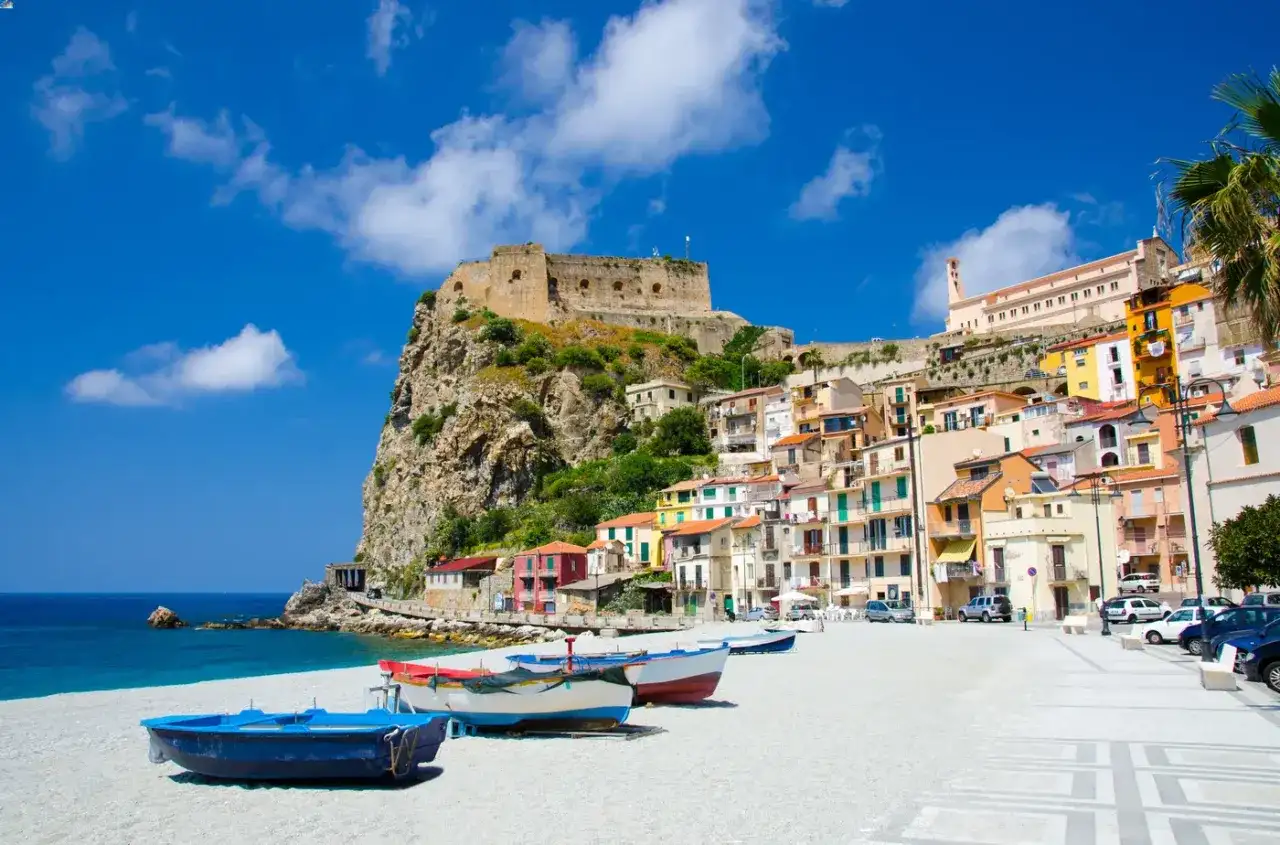 Vue d'une ville côtière italienne avec un château sur une falaise, des maisons colorées et des bateaux sur une plage.