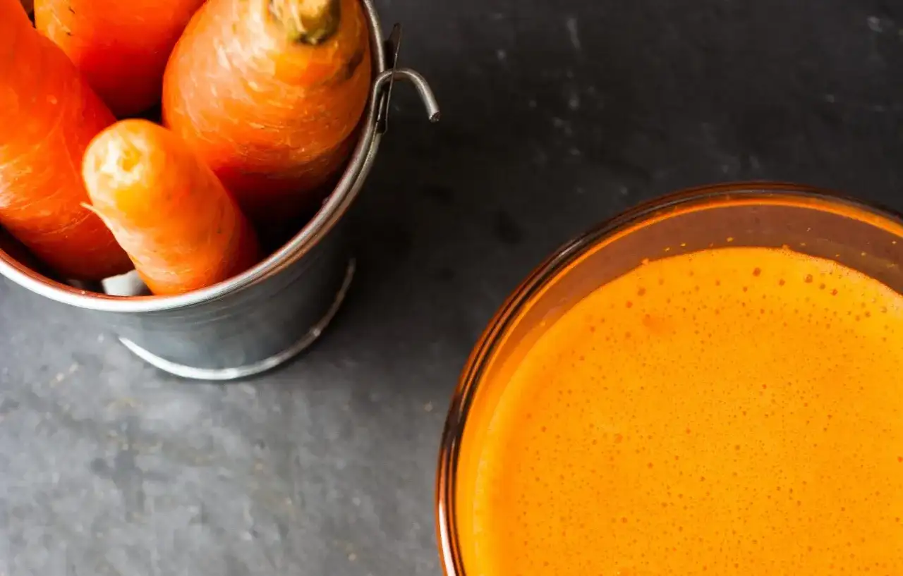 Frische Karotten in einem Eimer neben einem Glas leuchtend orangem Karottensaft. Die Wirkung von Karottensaft ist bekannt für seine Vitamine.