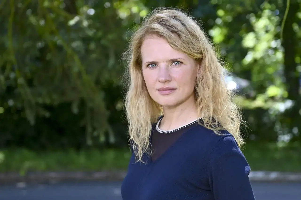 Wiktoria Grodecka z falowanymi blond włosami i niebieskimi oczami, w granatowym swetrze, na tle zieleni.