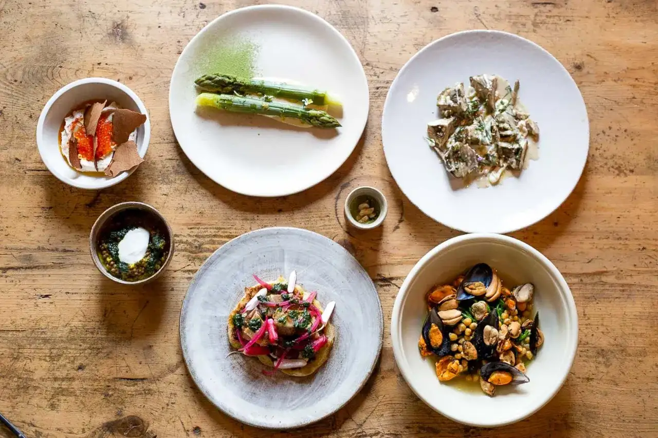Assortiment de plats gastronomiques : asperges, moules, tartare, et autres mets fins sur table en bois.