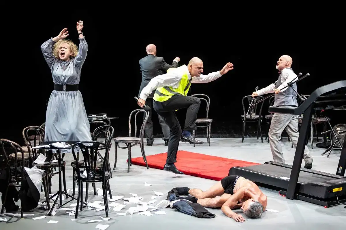 Scena teatralna z kobietą w szarej sukni, mężczyzną w kamizelce odblaskowej i leżącym na bieżni. Czy to symbol tego, ile trwają przerwy w teatrze?