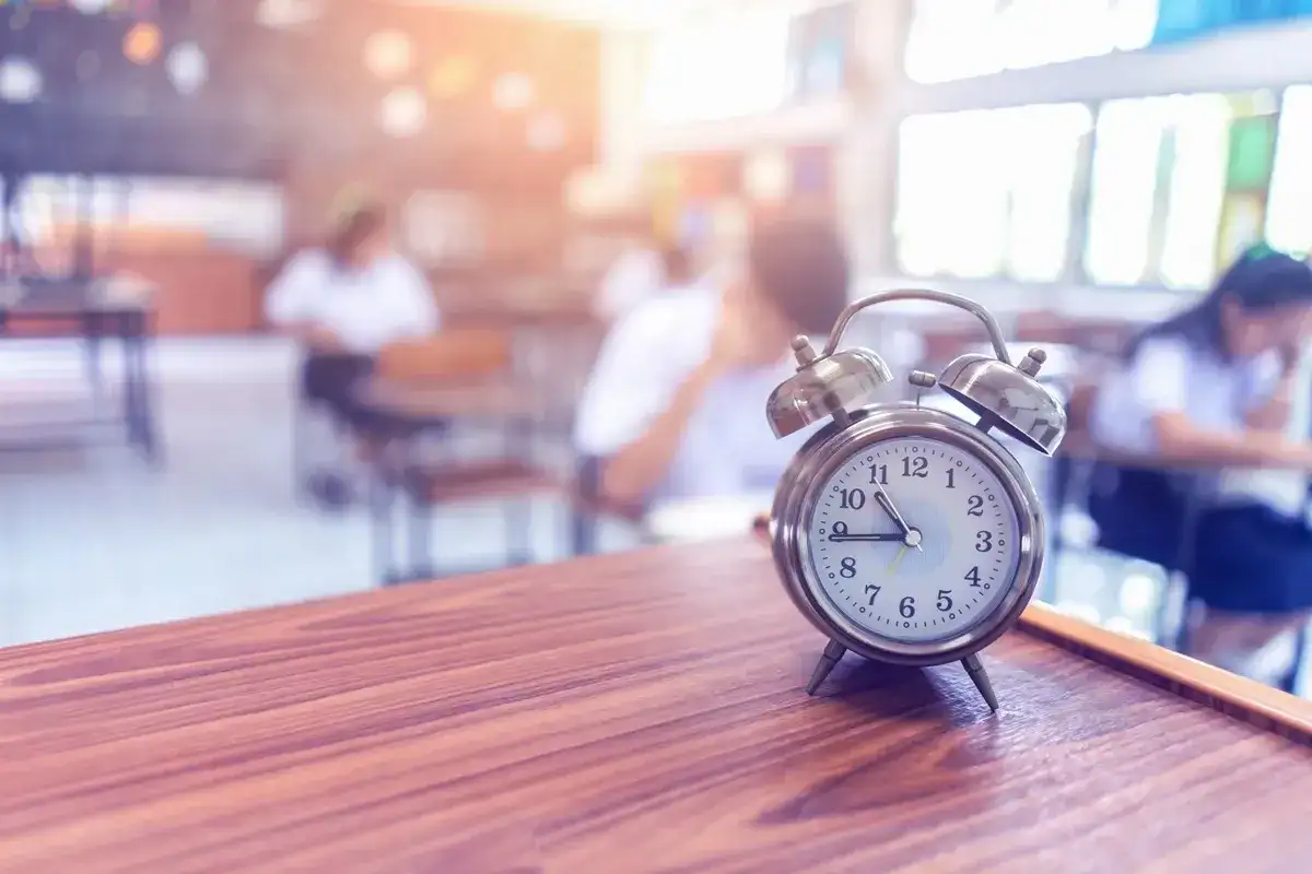 Budzik na biurku odmierza czas. Zastanawiasz się, ile trwa egzamin ósmoklasisty? Uczniowie w tle piszą test.