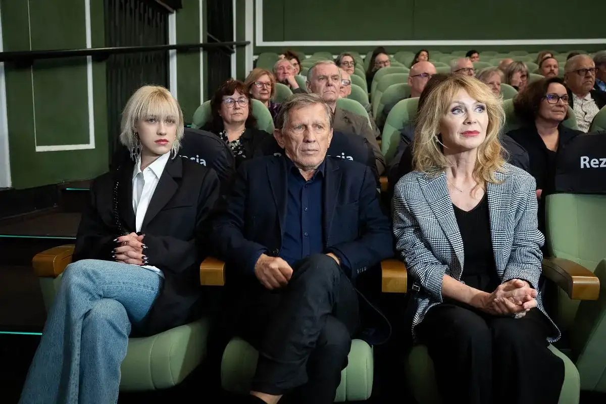Małgorzata Englert, w wieku, siedzi w pierwszym rzędzie obok innych osób, wpatrując się w dal.