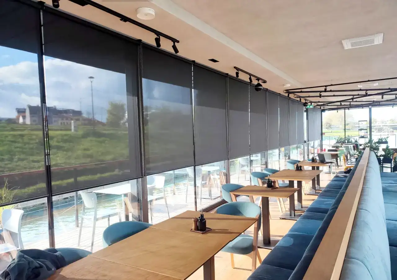 Nowoczesne wnętrze restauracji z widokiem na wodę, osłonięte eleganckimi roletami termicznymi na wymiar.