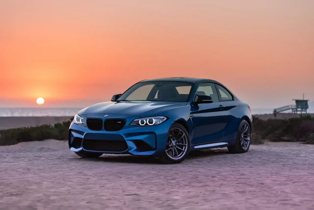 Niebieskie BMW M2 z pakietem M na plaży o zachodzie słońca. To jest m pakiet, który nadaje autu sportowy wygląd i osiągi.