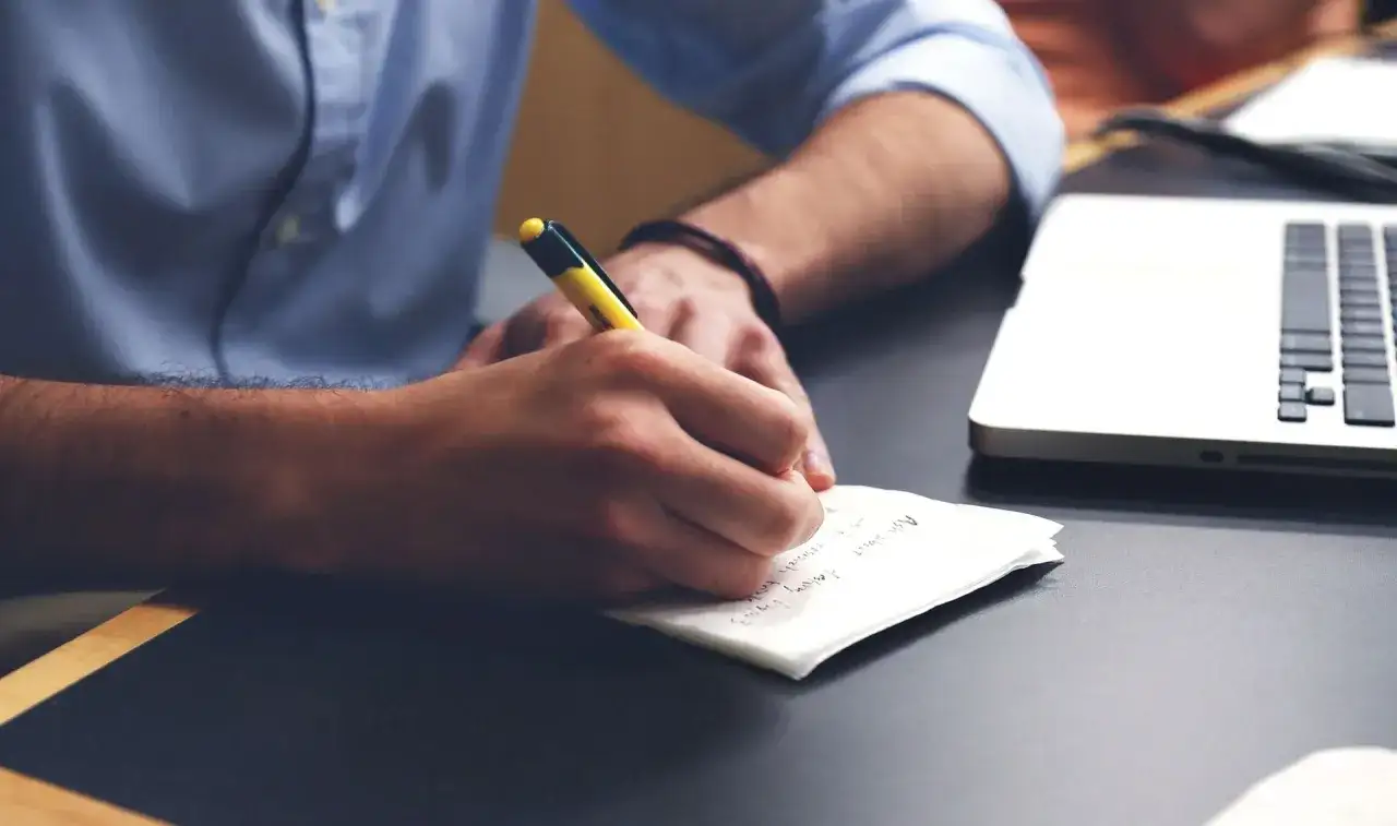 Mężczyzna w niebieskiej koszuli pisze coś długopisem na kartce. Obok leży laptop. To może być korekta świadectwa pracy.
