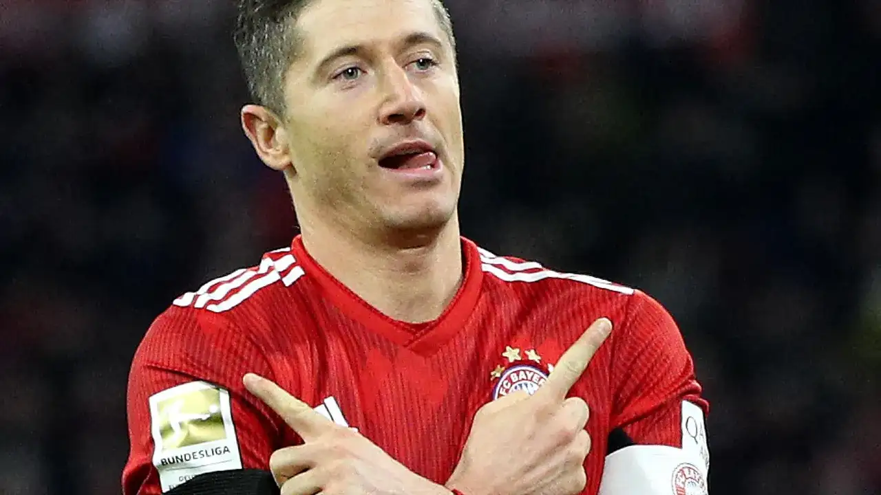 Robert Lewandowski w czerwonej koszulce Bayernu z naszywką Bundesligi, wykonuje gest radości po strzelonej bramce.