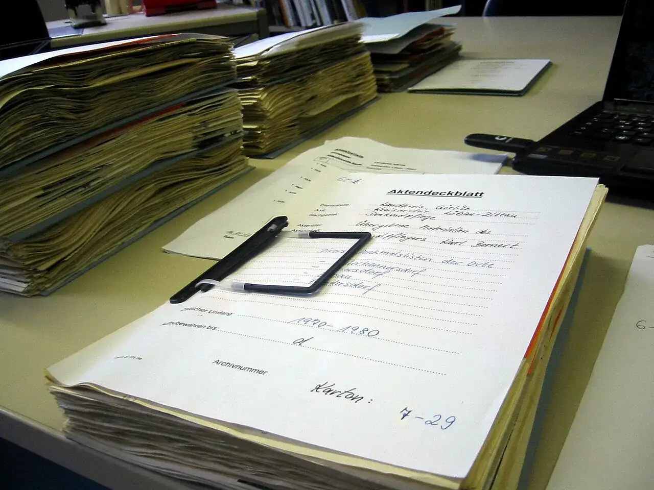 Stosy dokumentów, długopis i laptop na biurku. Na jednym z dokumentów widnieje napis "Akten-dossier co to", sugerujący porządkowanie akt.