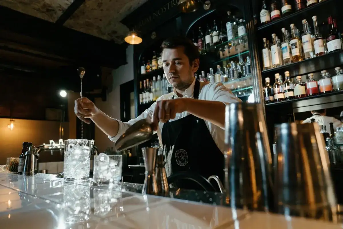Barman przygotowuje drinka, zastanawiając się, ile zarabia dobry barman. Za nim półki z alkoholem.