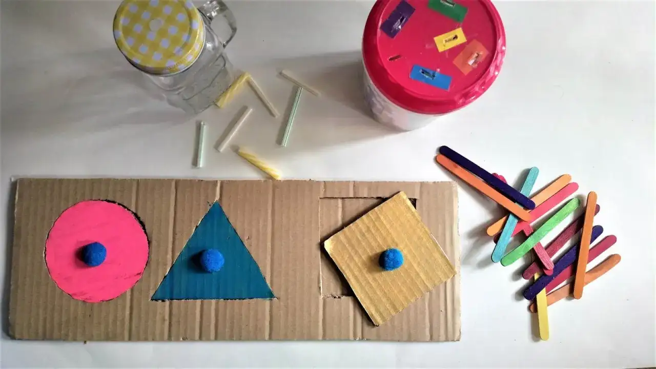 Kolorowe zabawki DIY dla niemowlaka: sorter kształtów z kartonu, patyczki i słoik.