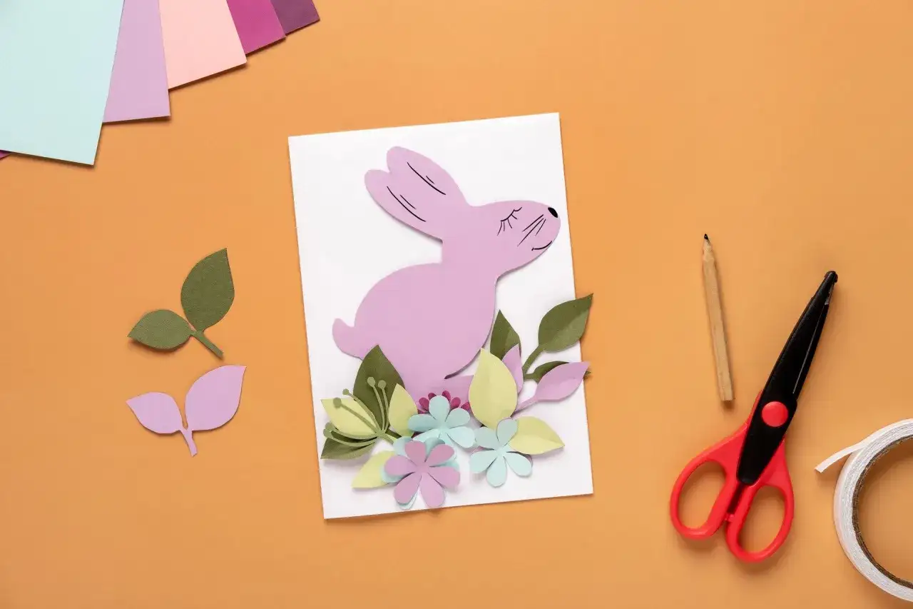 Uroczy różowy króliczek z papieru na kartce wielkanocnej DIY, otoczony kolorowymi kwiatami i liśćmi. Obok nożyczki, ołówek i taśma.