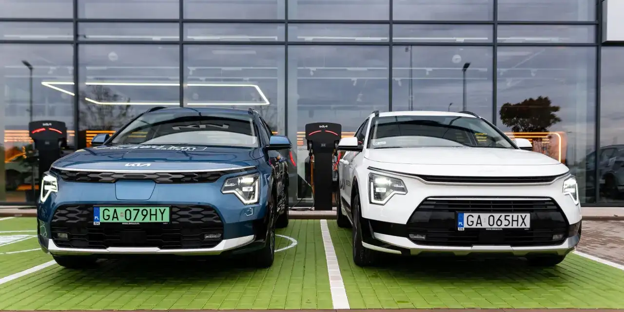 Dwa nowe samochody Kia Niro, jeden niebieski, drugi biały, stoją obok siebie na zielonym parkingu przed nowoczesnym budynkiem z dużymi oknami.