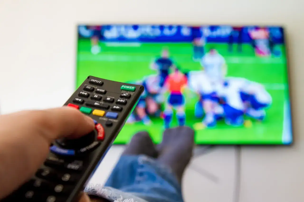 Dłoń trzymająca pilot do telewizora, na ekranie mecz piłkarski.