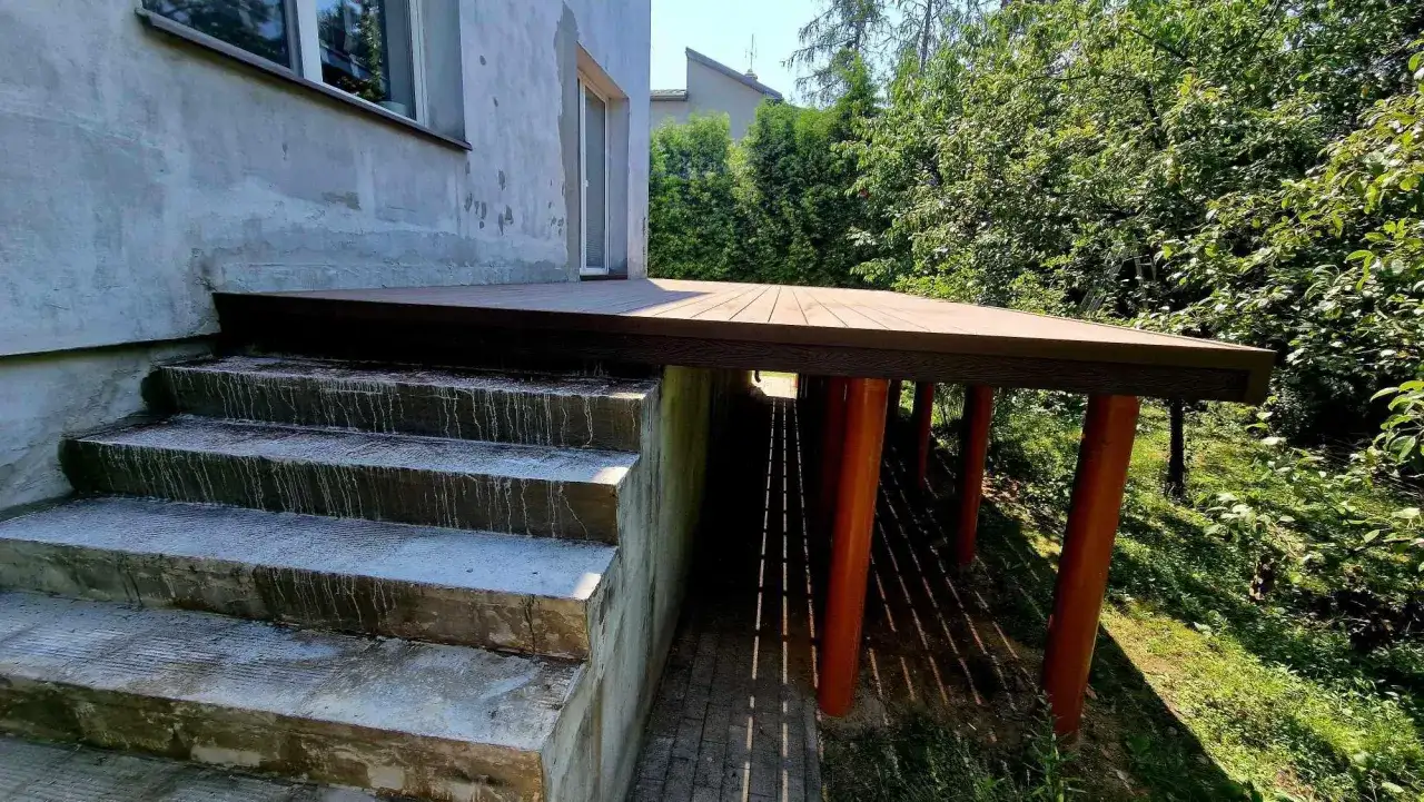 Nowy, wysoki taras na słupach, z drewnianą podłogą, sąsiaduje z betonowymi schodami i zielonym ogrodem.