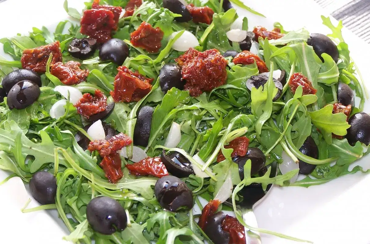Sałatka z rukolą, czarnymi oliwkami, suszonymi pomidorami i kawałkami mozzarelli, polana dressingiem.