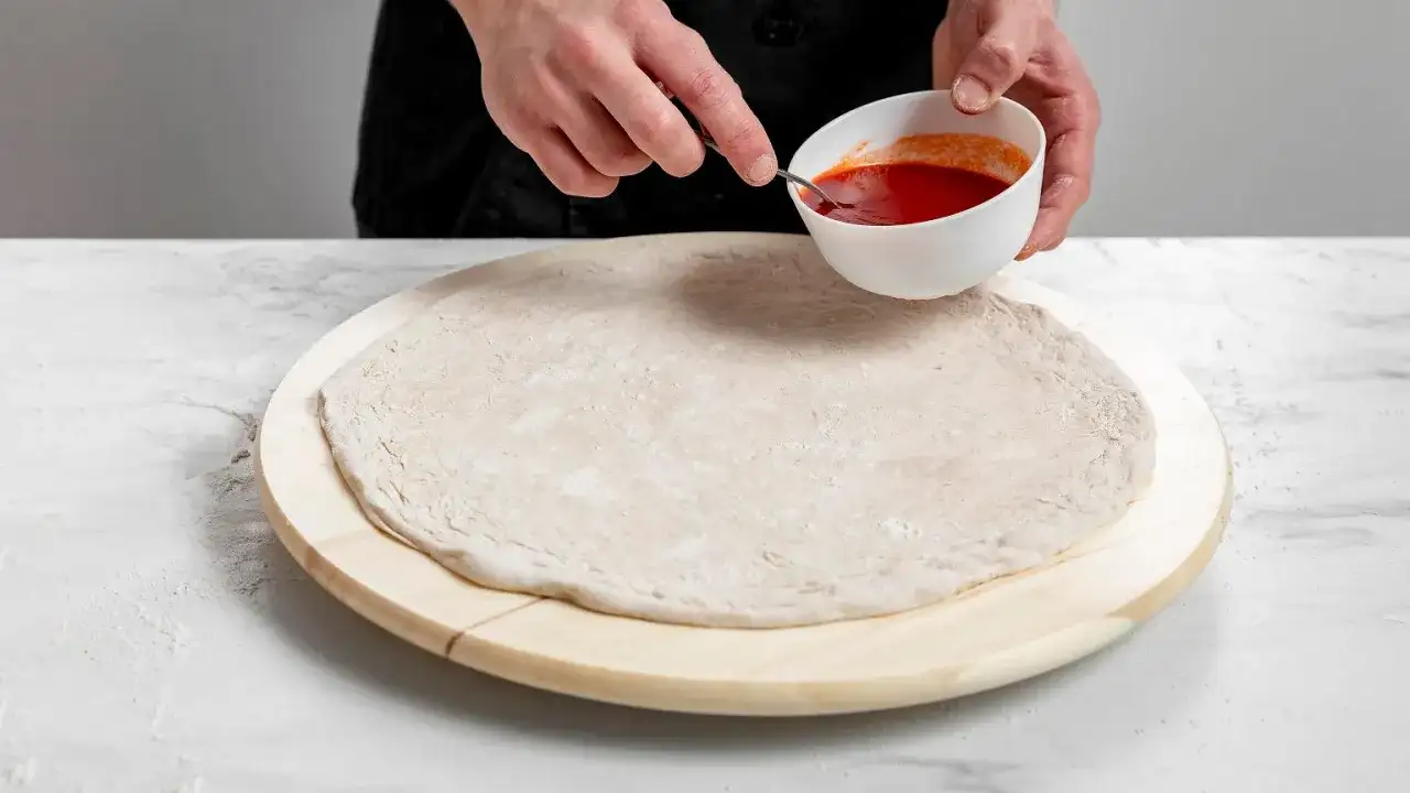 Ręce dodają sos pomidorowy do surowego ciasta na pizzę na drewnianej desce.