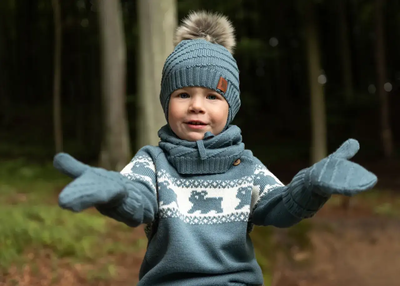 Uśmiechnięty chłopiec w zimowej czapce i rękawiczkach, ubrany w urocze sweterki dla dzieci z motywem renifera.