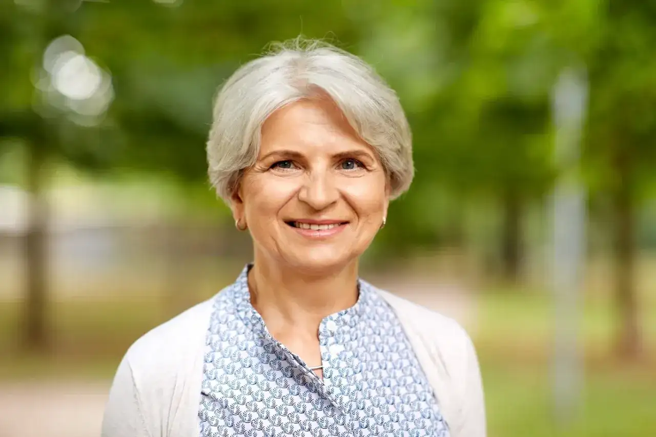 Uśmiechnięta starsza kobieta z siwymi włosami na tle zieleni.