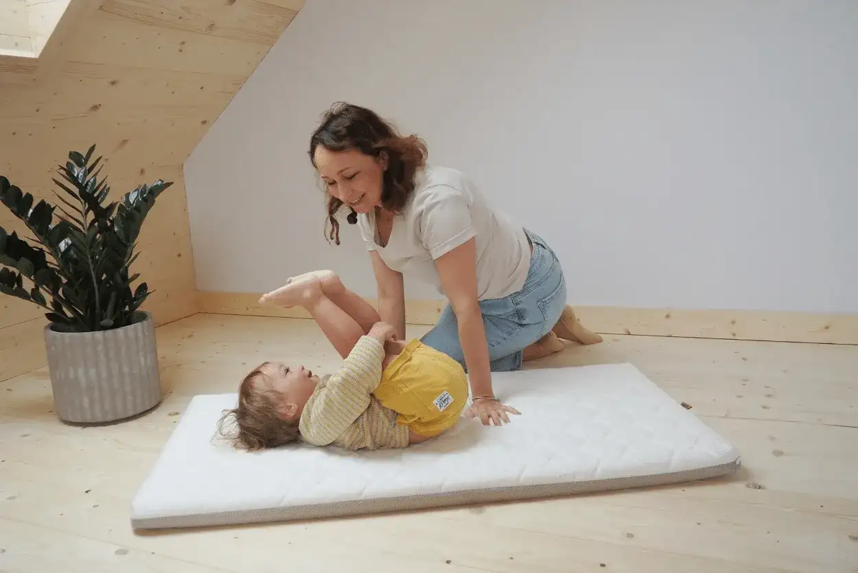 Mama bawi się z dzieckiem na materacu. Czy do łóżeczka turystycznego potrzebny jest materac? Tak, dla komfortu malucha.