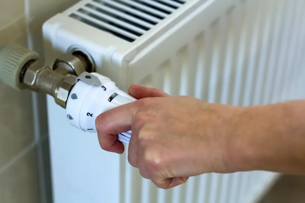 Dłoń reguluje termostat grzejnika, ustawiając temperaturę zgodnie z przepisami dotyczącymi minimalnej temperatury w mieszkaniu.
