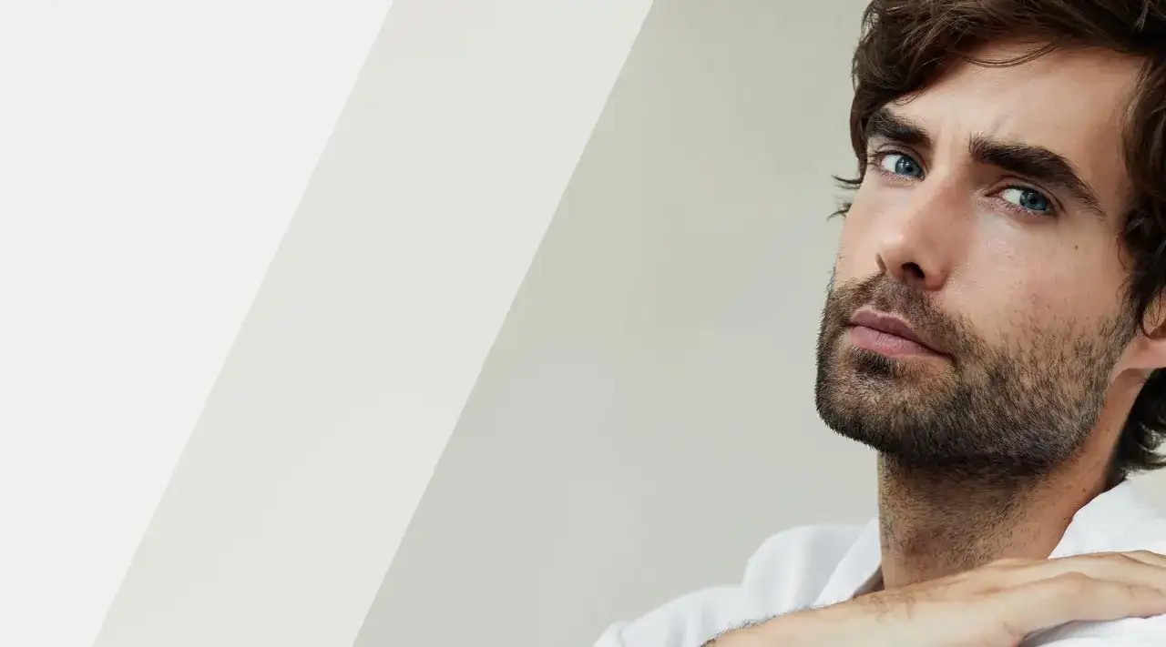 Mężczyzna z niebieskimi oczami i zarostem, zastanawiający się nad wyborem: zarost czy bez.