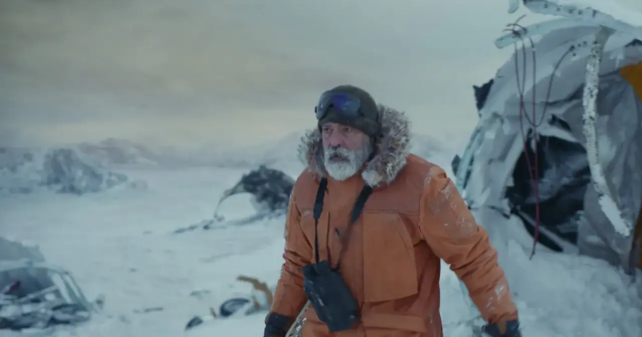 Samotny mężczyzna w pomarańczowej kurtce i goglach przemierza śnieżny krajobraz po katastrofie. W oddali widać szczątki samolotu, a nad wszystkim rozciąga się ponure niebo o północy.