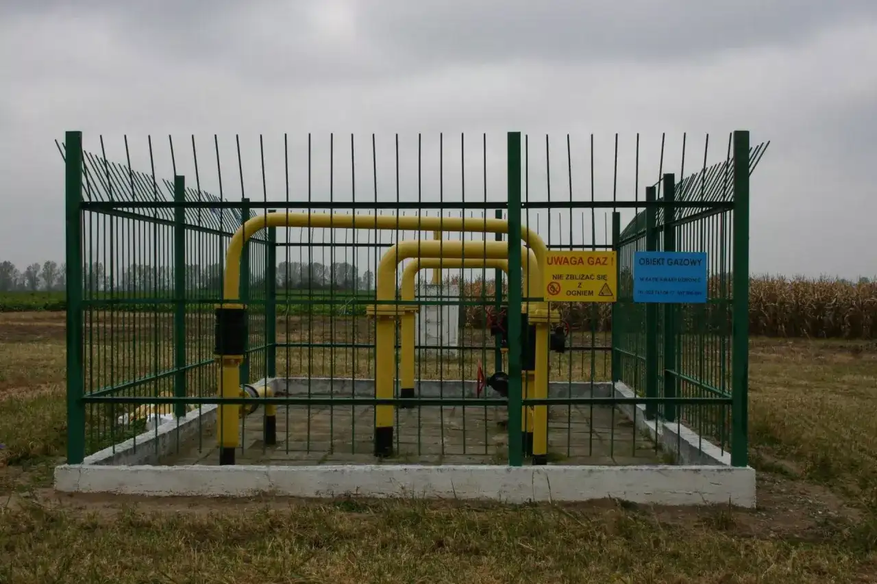 Ogrodzony obiekt gazowy z żółtymi rurami i tablicami ostrzegawczymi. Widać pola uprawne w tle. Określenie odległości budynku od gazociągu średniego ciśnienia jest kluczowe dla bezpieczeństwa.