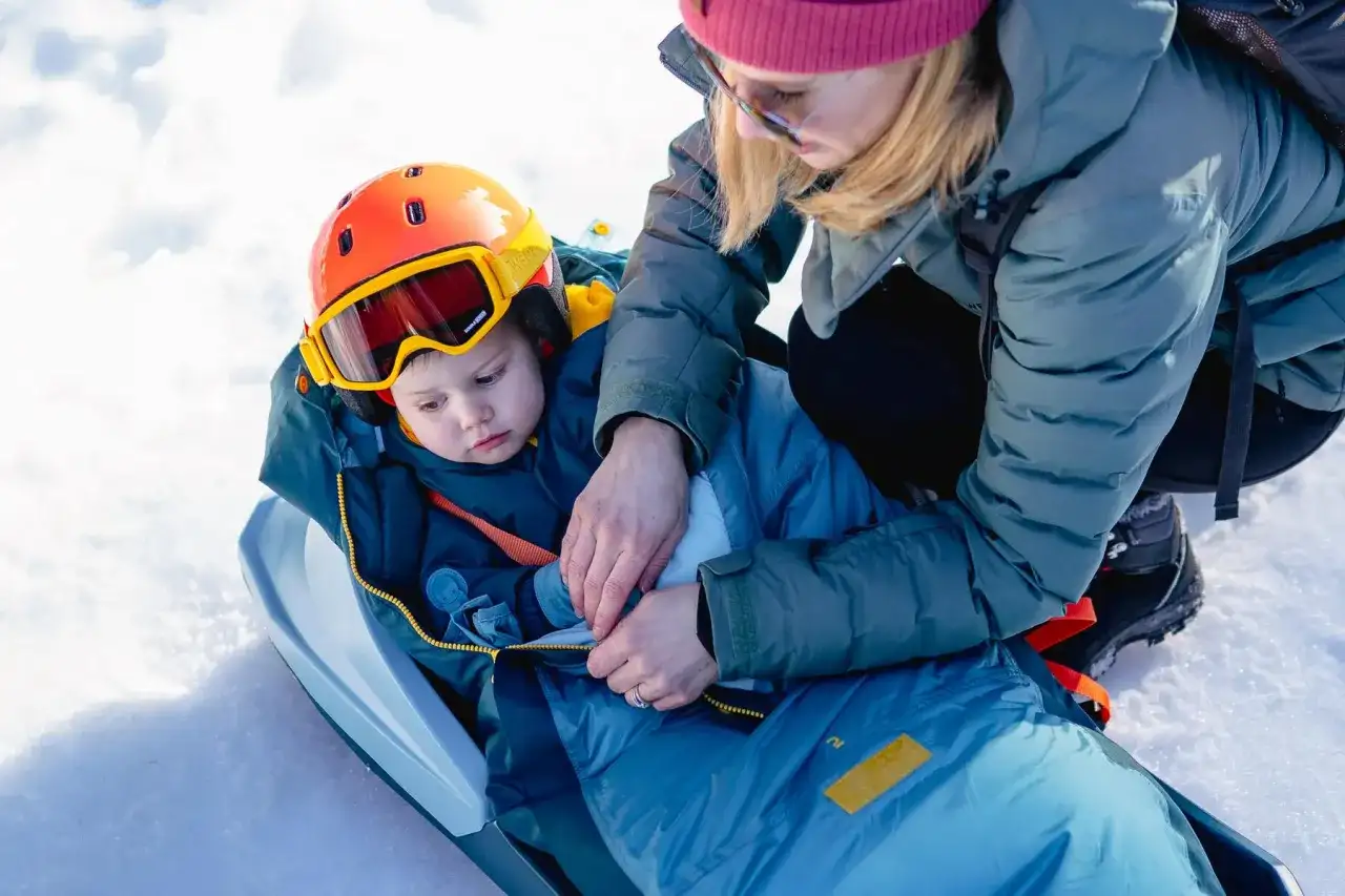 Mama pomaga dziecku ubrać się na sanki, zapinając śpiworek. Maluch ma kask i gogle narciarskie.