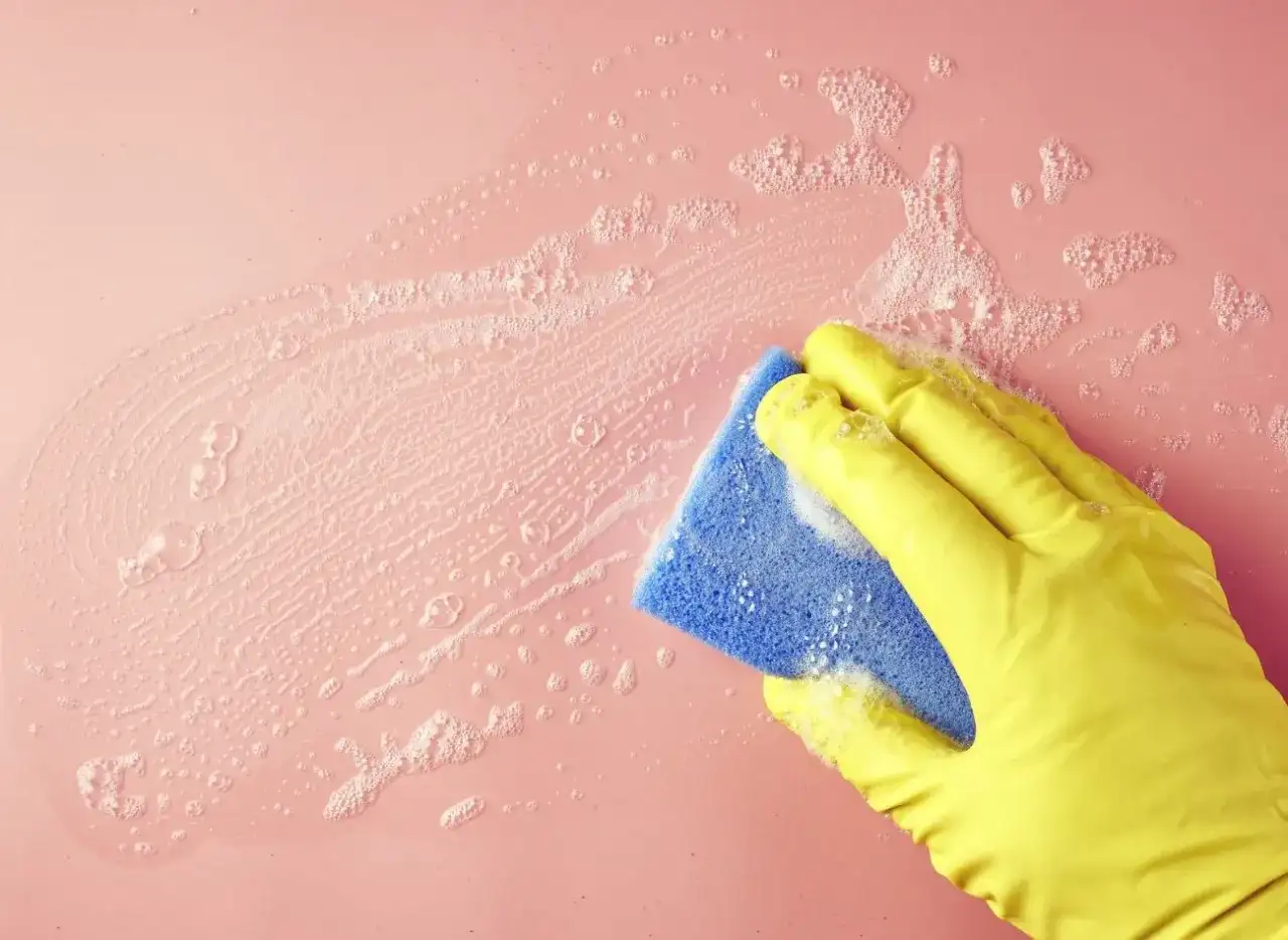 Dłoń w żółtej rękawiczce czyści różową ścianę niebieską gąbką z pianą.